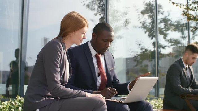 Two young businesspeople, black man and Caucasian woman, sitting on bench outdoors and discussing work using laptop computer, businessman using smartphone in the background