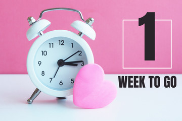 A small white alarm clock and a pink heart figurine. The inscription "1 week to go"...