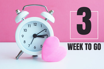 A small white alarm clock and a pink heart figurine. The inscription "3 week to go"...