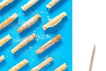 Dynamic composition with homemade cakes. Delicious waffle tubes with powdered sugar on a bright blue background. Delicious sweet snack, top view with copyspace to write recipe and pencil