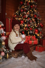 cute beautiful girl in a white sweater and red hat having fun and playing snowballs in winter on the street against the background of a wooden house. nearby are Christmas trees with lights