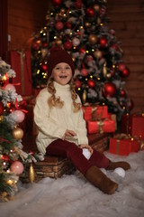 cute beautiful girl in a white sweater and red hat having fun and playing snowballs in winter on the street against the background of a wooden house. nearby are Christmas trees with lights