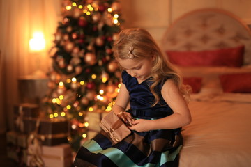 A cute girl with blond hair with a gift in her hands is waiting for the holiday against the background of the Christmas tree in the lights.