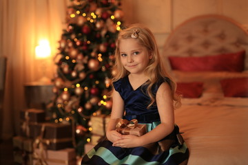 A cute girl with blond hair with a gift in her hands is waiting for the holiday against the background of the Christmas tree in the lights.