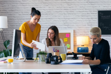 Three modern businesswomen talking and reviewing the latest work done on the digital tablet in a joint workspace.
