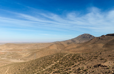 Overlooking a mountain range of Atlas Moutnain Range in Morocco. Some small bushes and trees seen in front of a lovely blue sky covered by some clouds.
