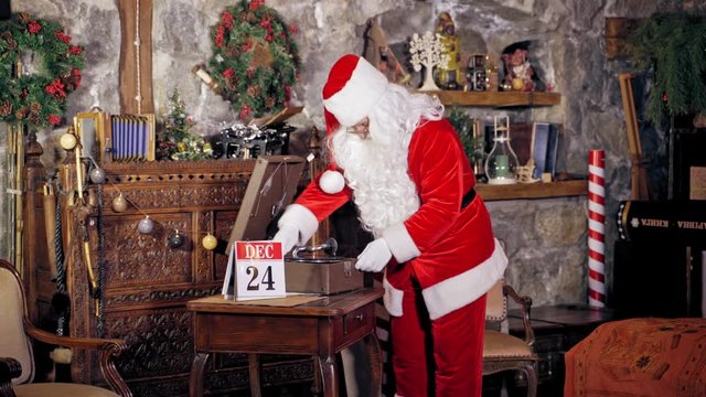 Santa Claus standing in specially decorated room, putting shellac or record on a gramophone or record player. Christmas spirit concept close up portrait. Christmas eve. New Year is coming