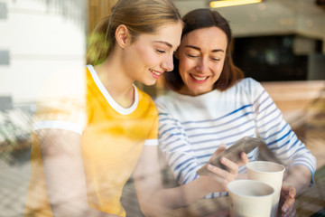 smiling mother and daughter sitting at cafe together looking at mobile phone