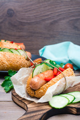 Appetizing hot dog made from fried sausage, rolls and fresh vegetables, wrapped in parchment paper on a cutting board on a wooden table