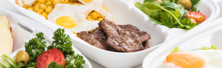 panoramic shot of eco package with corn, meat, fried eggs and salads on white background