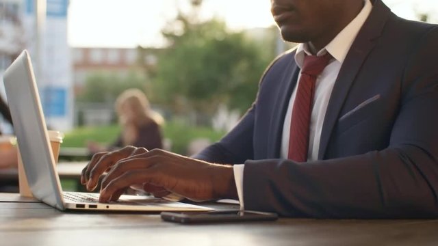 Medium shot of young black male entrepreneur in formal clothes concentrating on work and using laptop computer in outdoor coworking space in summer