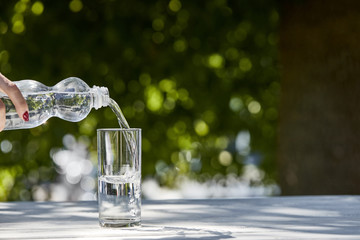 cropped view of woman pouring fresh clean water from bottle in transparent glass at sunny day on wooden table