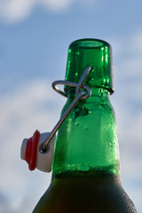 A selective-focus macro image of condensation - glistening enticingly in the afternoon sunshine - on a green-glass swing-top beer bottle; with a lightly clouded blue sky, blurry background.