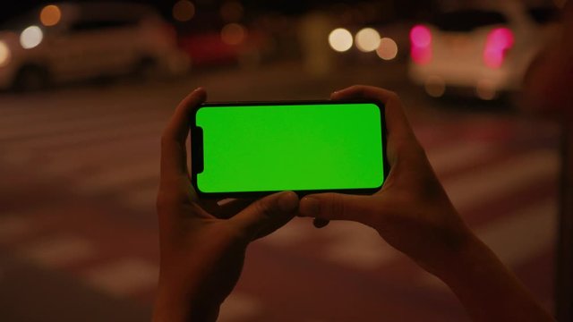 Handheld shot of young woman holding horizontal smartphone watching green screen mock-up device standing at traffic road in the evening. Urban life and technology.