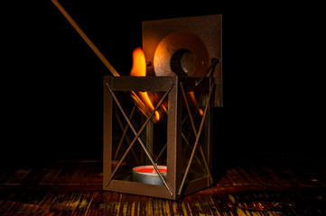 Glazed metal lantern with a red, flat candle. The candle is just lit with a long, burning match.