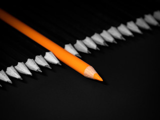 Black and white picture of the pencils with one orange coloured pecncil in the middle on a black backgound. Not like the others, being different.