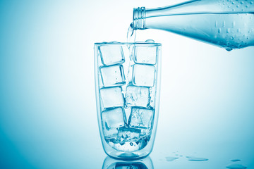 Pouring water from a glass bottle into glass with ice cubes on blue background