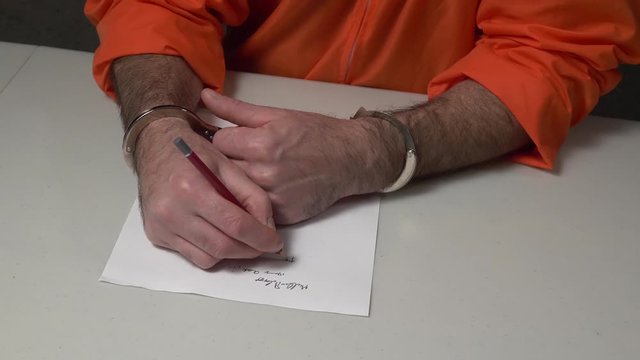 Prisoner dressed in a typical orange prison uniform trying to write something on a paper during his confession of a crime.