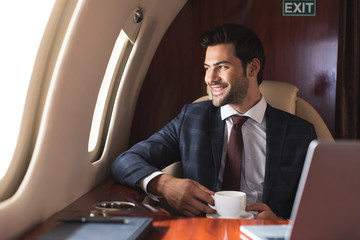 smiling businessman holding cup of coffee in plane during business trip - obrazy, fototapety, plakaty