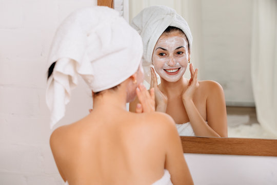 Woman With Towel Is Applying Mask On Her Face In Front Of The Mirror At Home. Skin Care.