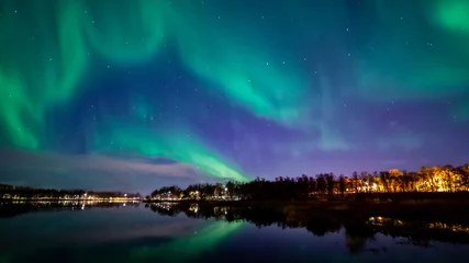 Foto op Plexiglas Northern lights above lake. Green aurora on purple sky with stars and clouds. Trees, city light. Reflections in water. Prestvannet, Tromso, Norway. © Ida Haugaard Olsen