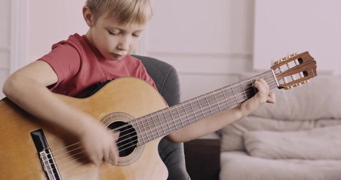 Young boy strumming a guitar. Boy of 8 years plays the classical guitar. The caucasian child learns to play the guitar. Happy child loves playing music. Real time. Zoom in. Natural lighting.