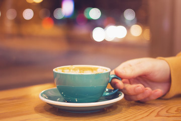 Man’s hand with cup of coffee in a cafe on a background of city lights behind window. Toned photo.