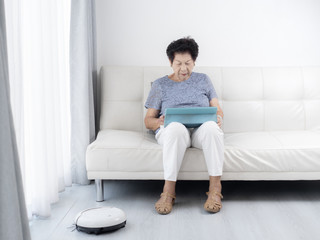Senior woman unsing tablet while robot vacuum cleaning floor at home. Modern lifestyle concept.