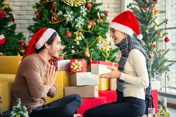 Obraz na płótnie Canvas Happy young adult man in santa hat give a Christmas present in gift box to beautiful woman girlfriend with Christmas tree decoration background. Christmas family celebration festive party concept.