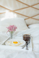 Fototapeta na wymiar White tray with breakfast on a bed in a hotel room. Fried egg, cup of coffee and flowers in white sheets in light bedroom.