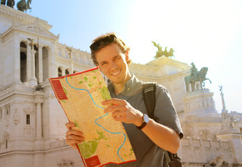 Handsome guy holding a city map looking at the camera and smiling at the beautiful sunny day at the Rome, Italy. The famous Vittorio Emanuele II monument at Piazza Venezia