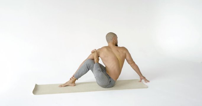 A flexible and plastic male yogi with a beard does yoga on a rug in a White Studio. He does twists and stretches.