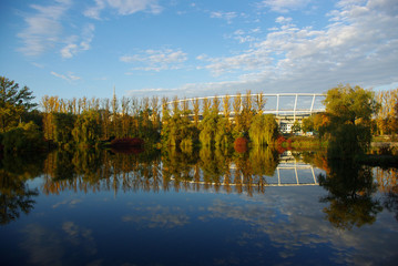 Obiekt Stadionu Śląskiego w Chorzowie odbijający się w parkowym stawie wraz z otaczającymi go drzewami pełnymi kolorowymi liśćmi wczesną słoneczną jesienią na tle błękitnego nieba.