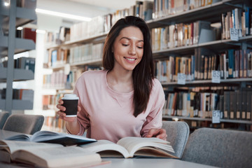 With plastic cup of coffee in hand. Brunette girl in casual clothes having good time in the library...