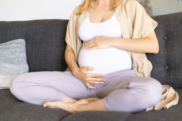 Expectant mother embracing baby bump. Pregnant woman sitting on couch, touching belly. Expecting baby concept