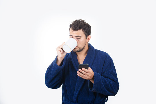 Portrait of sleepy young man messy hair, wears blue bathrobe, drinking coffee from his cup and speking on phone isolated on white background.