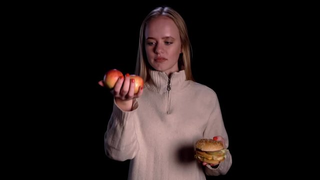 Pretty blondie woman in white sweater choosing between apple and burger over black background. Healthy food. Girl on a diet.