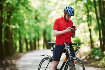 Using smartphone. Cyclist on a bike is on the asphalt road in the forest at sunny day