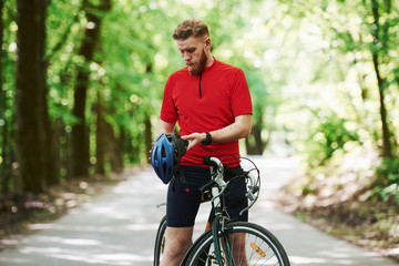 Fototapeta premium Taking off protective helmet. Cyclist on a bike is on the asphalt road in the forest at sunny day