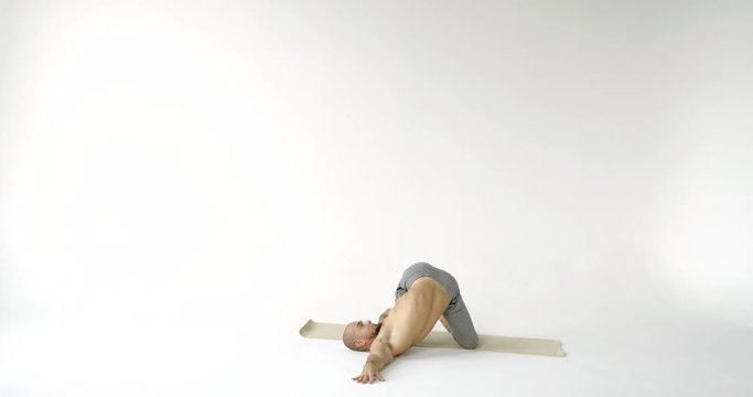 A flexible and plastic male yogi with a beard does yoga on a rug in a white Studio. He pulls the muscles of the sides and does a twist.