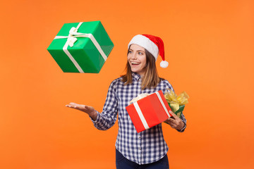 Portrait of happy brunette young woman in santa hat and checkered shirt standing throwing gift boxes in air or catching presents, christmas shopping. indoor studio shot isolated on orange background