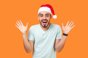 Fototapeta na wymiar Portrait of amazed happy bearded young man in santa claus hat and casual white t-shirt standing with open mouth and raised hands, excited and shocked. indoor studio shot isolated on orange background