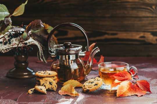 Autumn teatime composition on dark background with colored leaves and chocolate cookies, sun light beam on the cup behind the glass with water dew drops