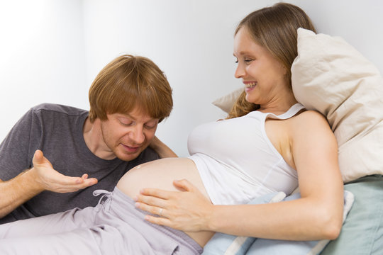 Happy expecting couple having fun at home. Future dad talking to pregnant wife tummy. Parenthood concept - Powered by Adobe