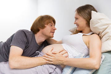 Obraz na płótnie Canvas Happy pregnant couple enjoying leisure time at home. Excited future dad embracing baby bump, smiling, looking at camera. Family and love concept