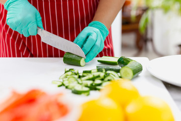 Woman with green glove cutting cucumber in the kitchen on chopping board.