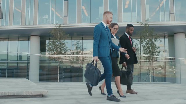 Male and female business people walk and discuss business. Group of colleagues going along modern glass business center. Three confident persons in formal suits