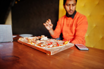 Confident young indian man in orange sweater sitting at pizzeria and eat pizza.