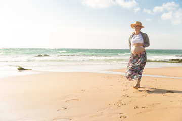 Fototapeta na wymiar Cheerful future mother walking on beach during vacation. Happy pregnant woman holding hand on belly. Pregnancy concept