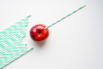 a ripe juicy bright red apple and in it a striped white and green paper drinking straw, and a pile of drinking straws nearby, located on a white background